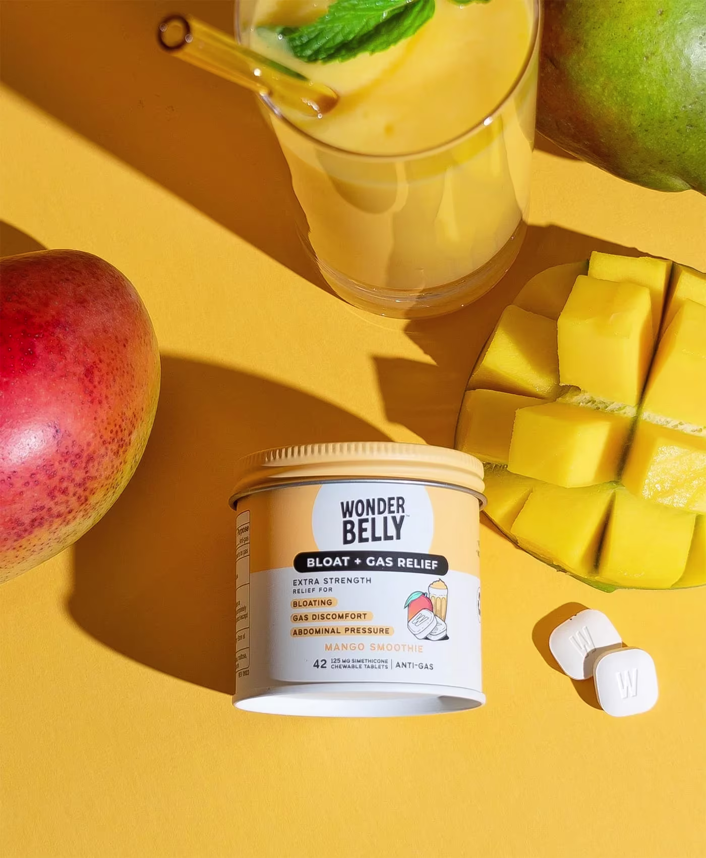 Wonderbelly bloat and gas relief jar labeled “Mango Smoothie” placed among fresh mango, a mango smoothie drink, and chewable tablets on a bright yellow background.