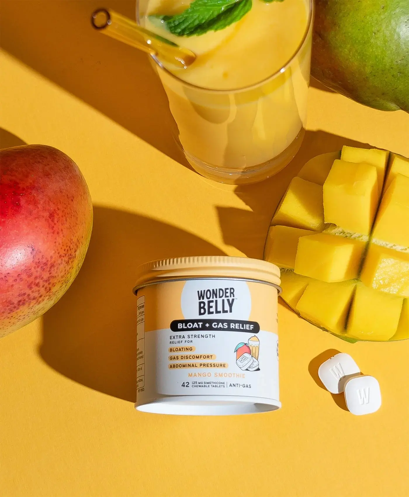 Wonderbelly bloat and gas relief jar labeled “Mango Smoothie” placed among fresh mango, a mango smoothie drink, and chewable tablets on a bright yellow background.