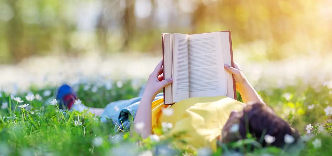 Child lying down on grass and reading a book in warm sunlight.