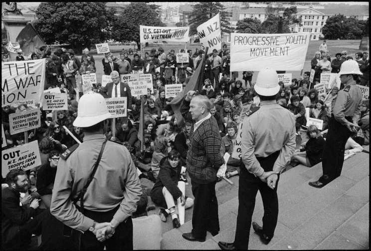 Protesters, with banners and signs, demanding the withdrawal of New Zealand and United States troops from Vietnam, Parliament, Wellington, 1969. Jim Hoy is making a speech from Parliament steps, flanked by two policemen.