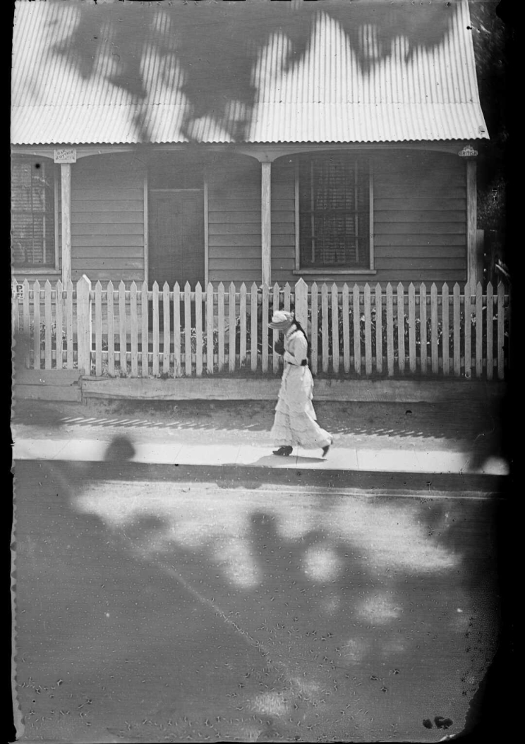 A woman walks on the footpath in front of white picket fence and a wooden house in the background, the ground is dappled in shade from a tree.