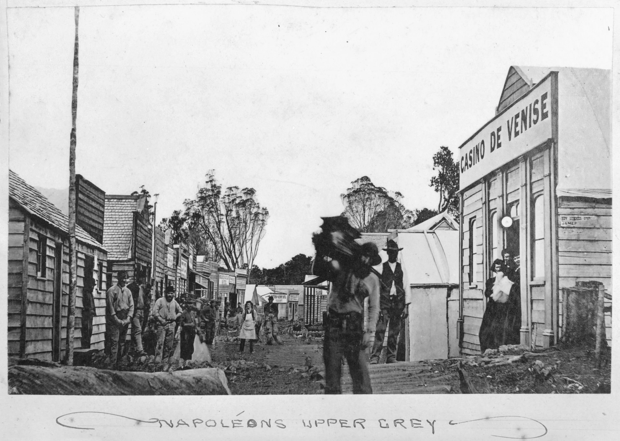 A street scene with men and women standing outside various buildings along a muddy street.