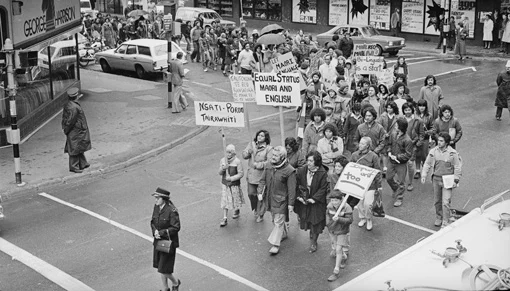 Black and white image of a group of protesters in 1980's clothing holding signs in support of equal status of the Māori language with English.