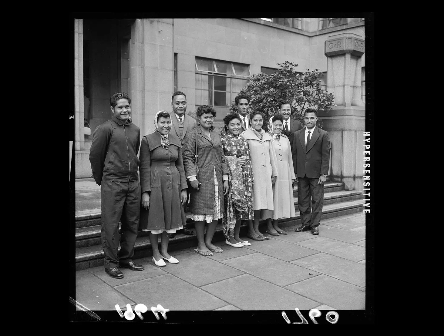Group of young men and women from the Tokelau Islands wearing coats and warm clothing standing in front of a building.