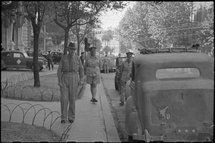 Street scene with men in khaki clothing standing besides cars parked against the kerb.