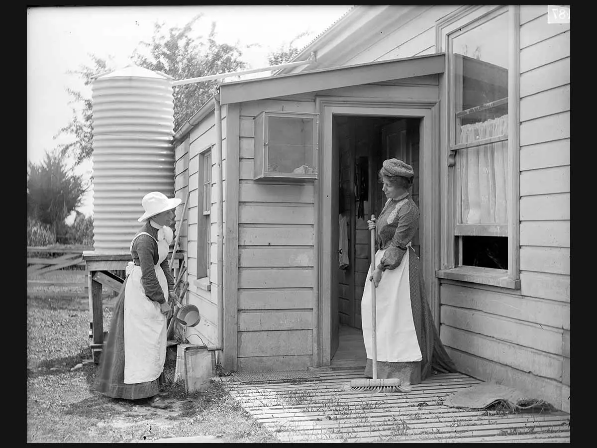 2 people standing outside a house doing housework. They are dressed in aprons and hats, standing by the back door of a house. They are sweeping with a broom and cleaning pans.