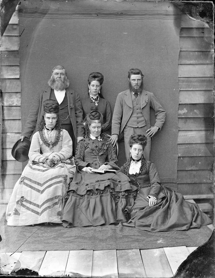 Victorian era photo of a family of six people posed in front of a blanket pinned to a wooden wall. 