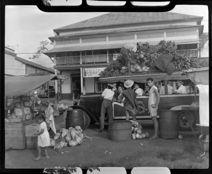 A black and white photo showing a fruit stand and truck with fruit and produce piled high on the roof. 