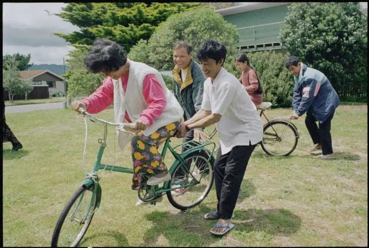 A scene on green grass of two people learning to ride bicycles while being assisted by two others. 