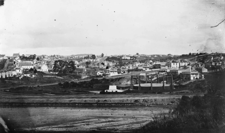 A black and white landscape photo showing the edge of town with dirt fields and houses in the distance.