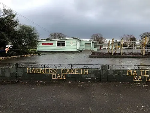 Colour photograph of a kuaka (bar-tailed godwit) cut out held in front of Takapau Primary School. The words 'Manners maketh man' are displayed on a fence.
