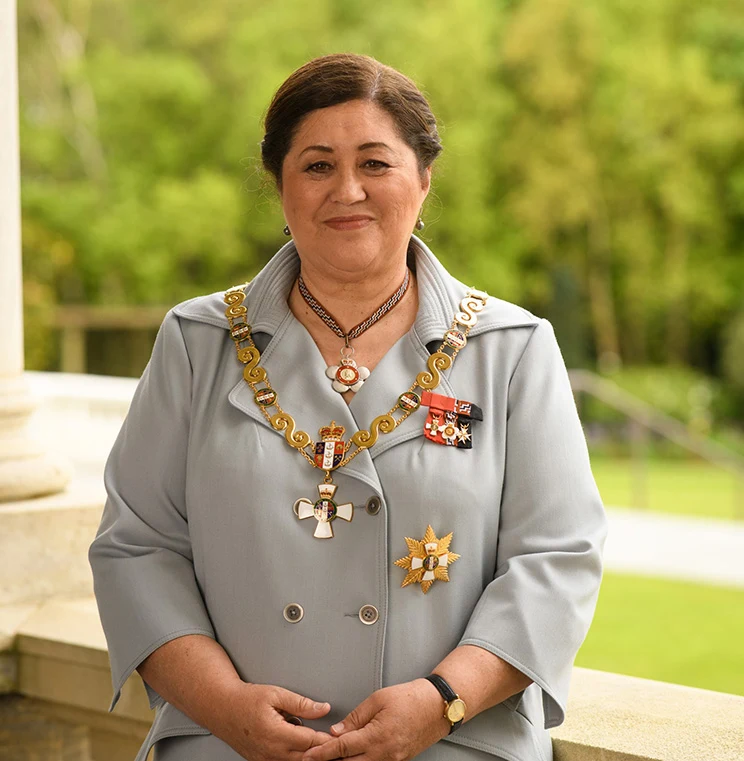 A Māori woman wearing a pale grey jacket, gold chains of office, and a number of medals.