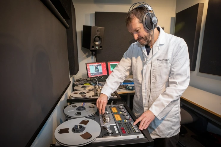 AV technician seen in white lab coat operating a large reel to reel tape machine while wearing headphones.