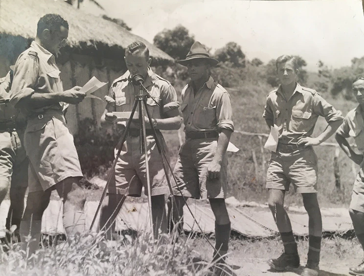 A man wearing army uniform is reading from a piece of paper into a microphone on a tripod, watched by four other soldiers.