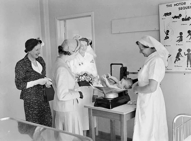 Two nurses and two women wearing skirt suits, hats and gloves smiling at a baby lying on baby scales set on a table.