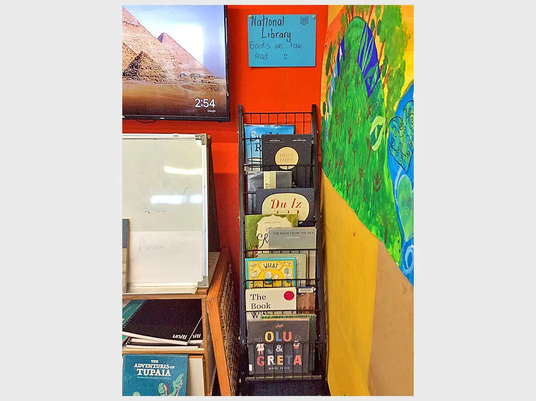A front-facing bookshelf in a classroom displaying picture books. Above the shelf is a handwritten sign with the words, 'National Library — Books we have read'.