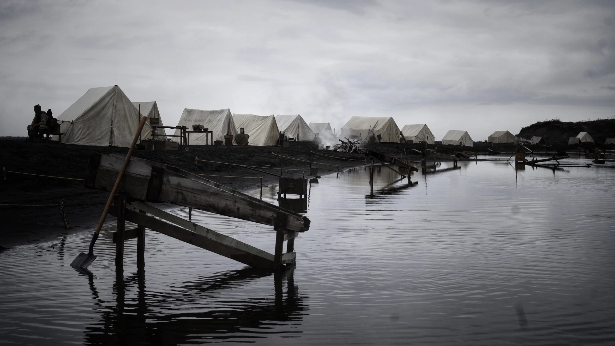 Tents line a river bank with wooden structures standing in the water. 