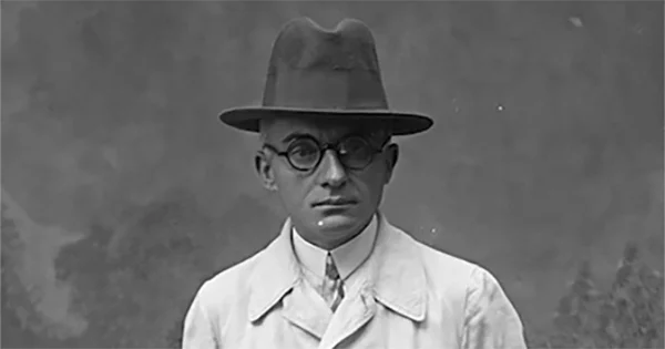 Head and shoulders image of a man wearing an overcoat, a homburg hat and round glasses.