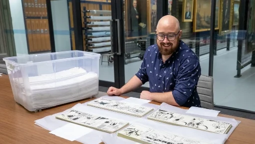 Man sat behind a table. On top of the table is a collection of comic strips.