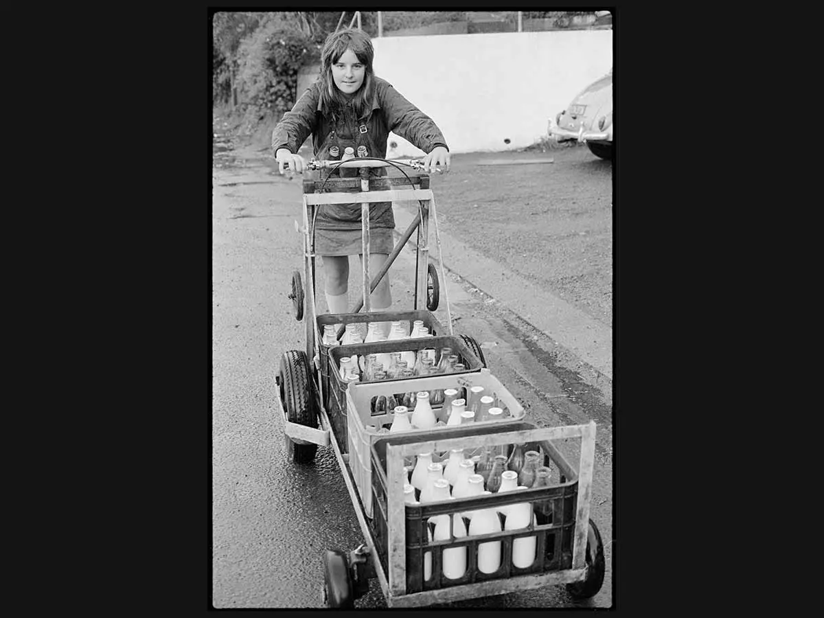 A young person is pushing a cart filled with crates of glass milk bottles along a footpath. They are wearing an apron with more milk bottles in their pocket.