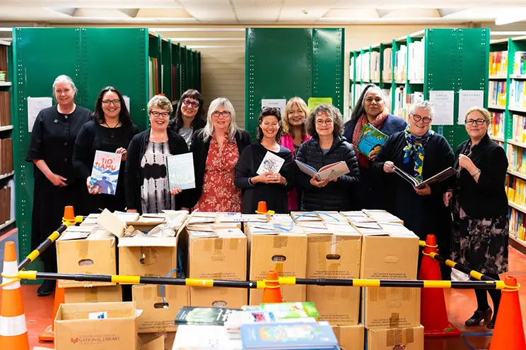 A group of women standing in front of bookshelves, holding books and there is a big pile of boxes in front of them.