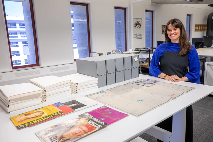 A woman stands behind a table on which sit magazine collections and archival folders and boxes.