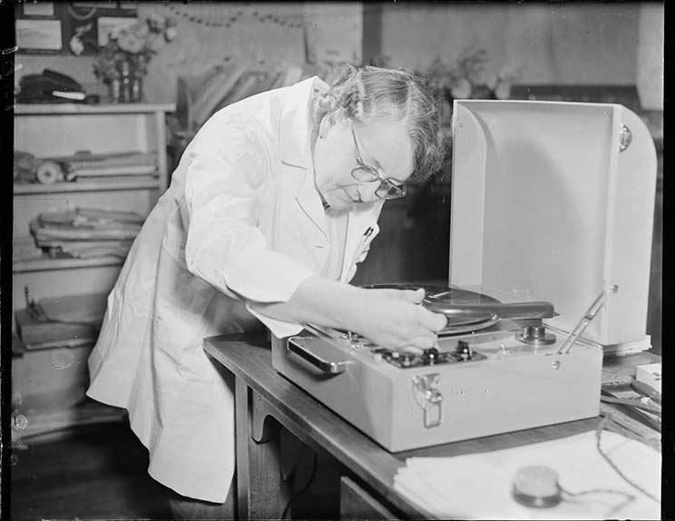 Woman in white coat moving the arm of a record player to start a record playing.