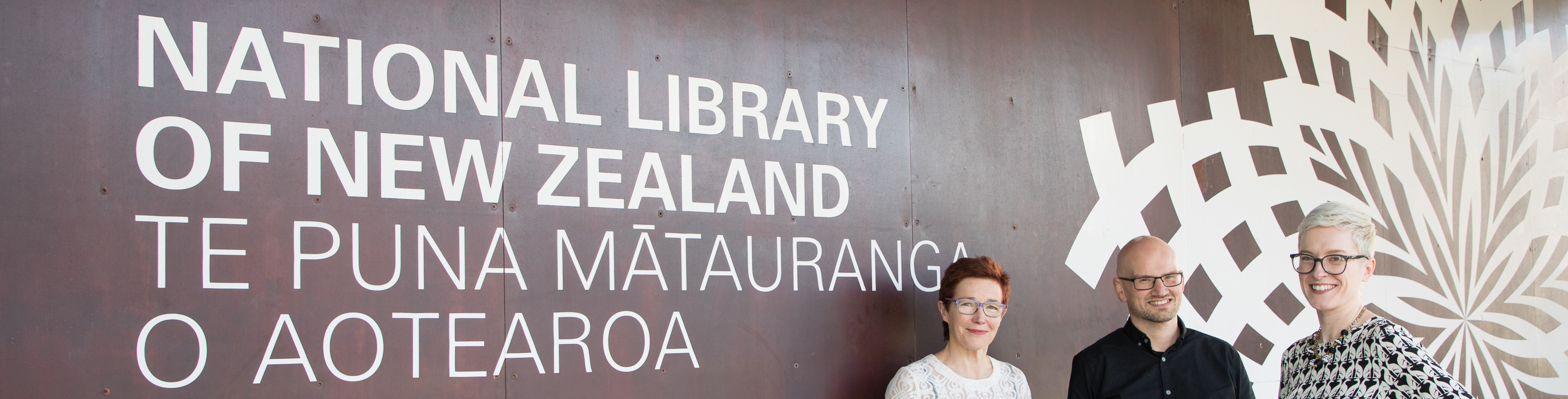 Two women and one man standing in front of a sign that reads National Library of New Zealand Te Puna Mātauranga o Aotearoa.
