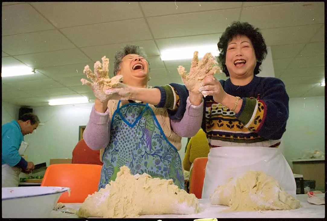 Ng Soon Yee (left) and Mel Jean Chin laughing and holding up their hands, covered in dumpling mixture, from the piles of mixture in front of them.