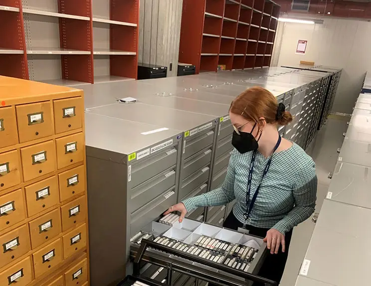 Woman in a room with lots of drawers looking at a trolley with cassette tapes on it.