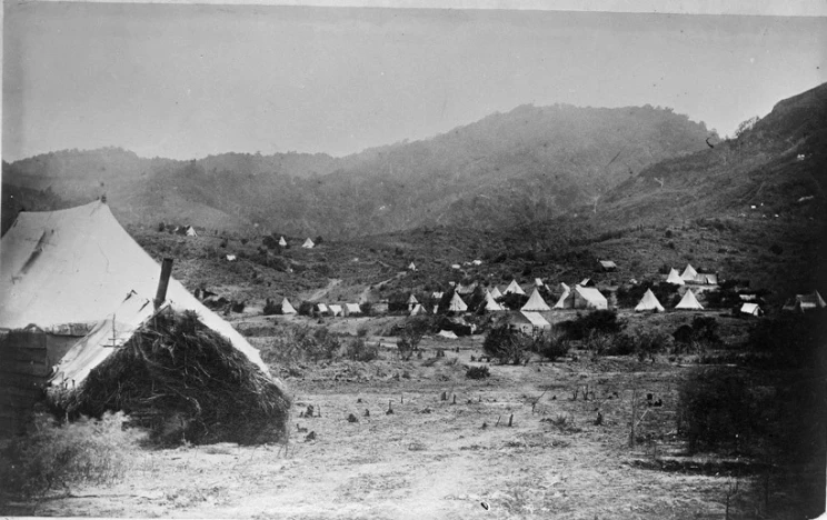 A black and white photograph showing numerous white tents scattered about the landscape with low, bush-clad hills in the background.