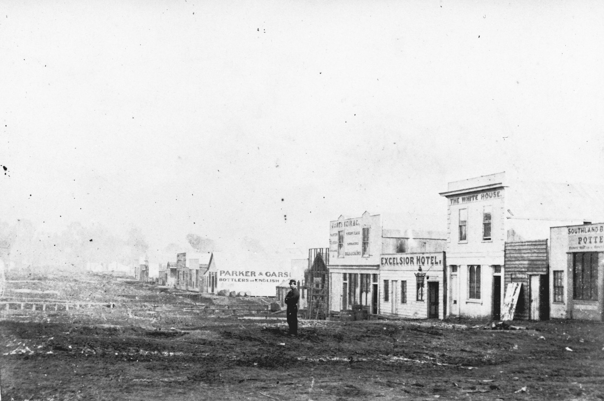 A lone man stands in a large, dirt street with wooden buildings on one side.