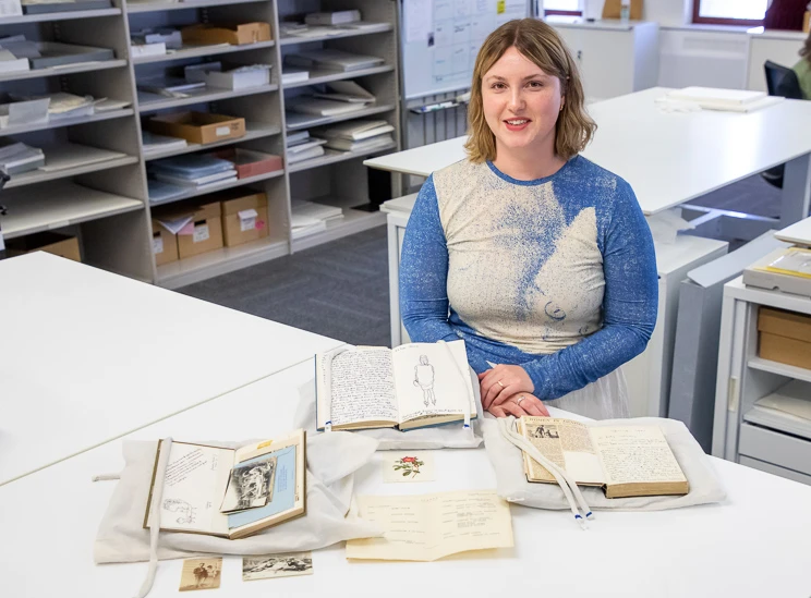 A woman seated behind a desk with various notebooks displayed on it, each held open with a string weight or snake weight.