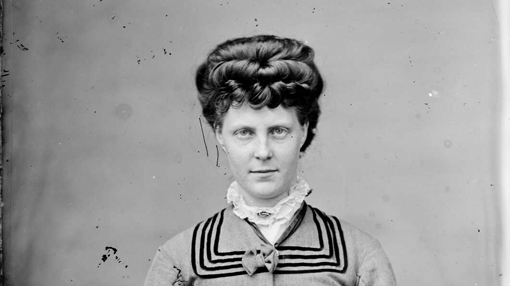 Black and white Victorian-era studio photograph of a woman wearing a jacket with a sailor boy collar, a matching skirt, and a ruffled high-neck blouse. She has a Mona Lisa smile.