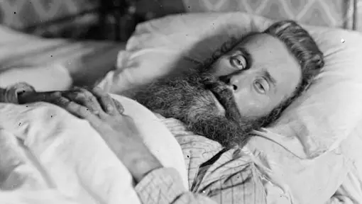 Victorian era black and white photo of a man lying in a bed looking at camera. He may be unwell. 