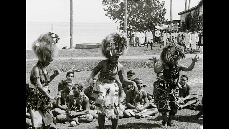 Three Samoan women performing a traditional dance with a crowd of people on the grass behind.