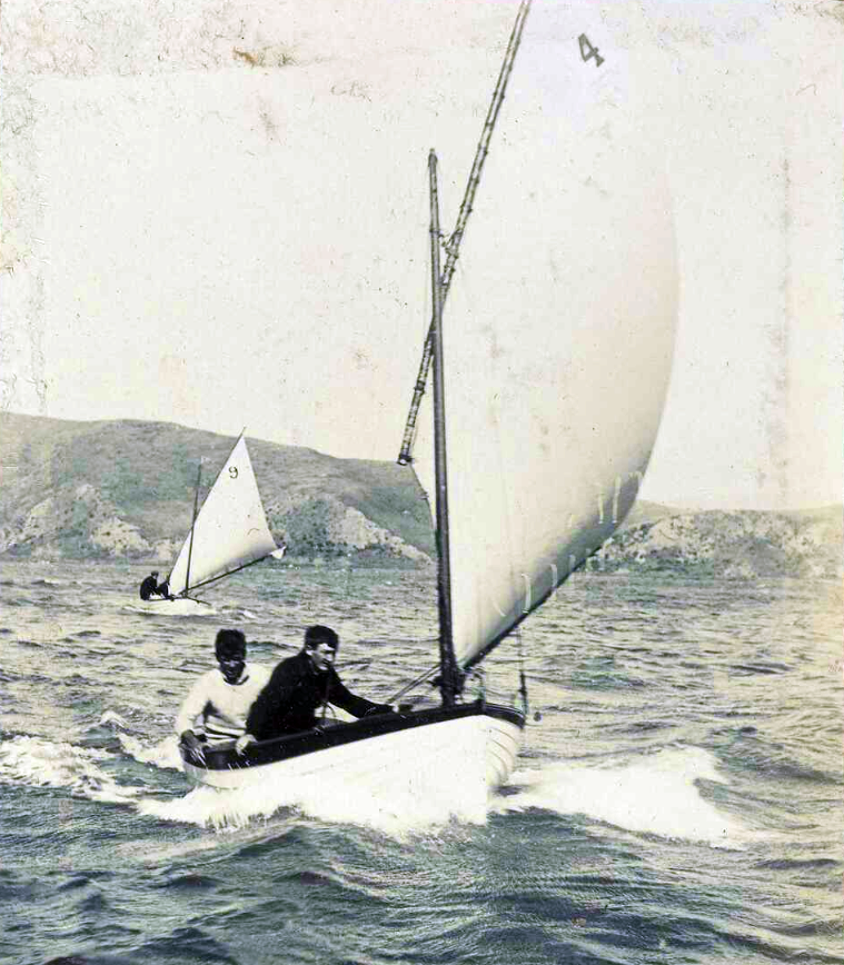 Shows a small boat under sail with two men on board.