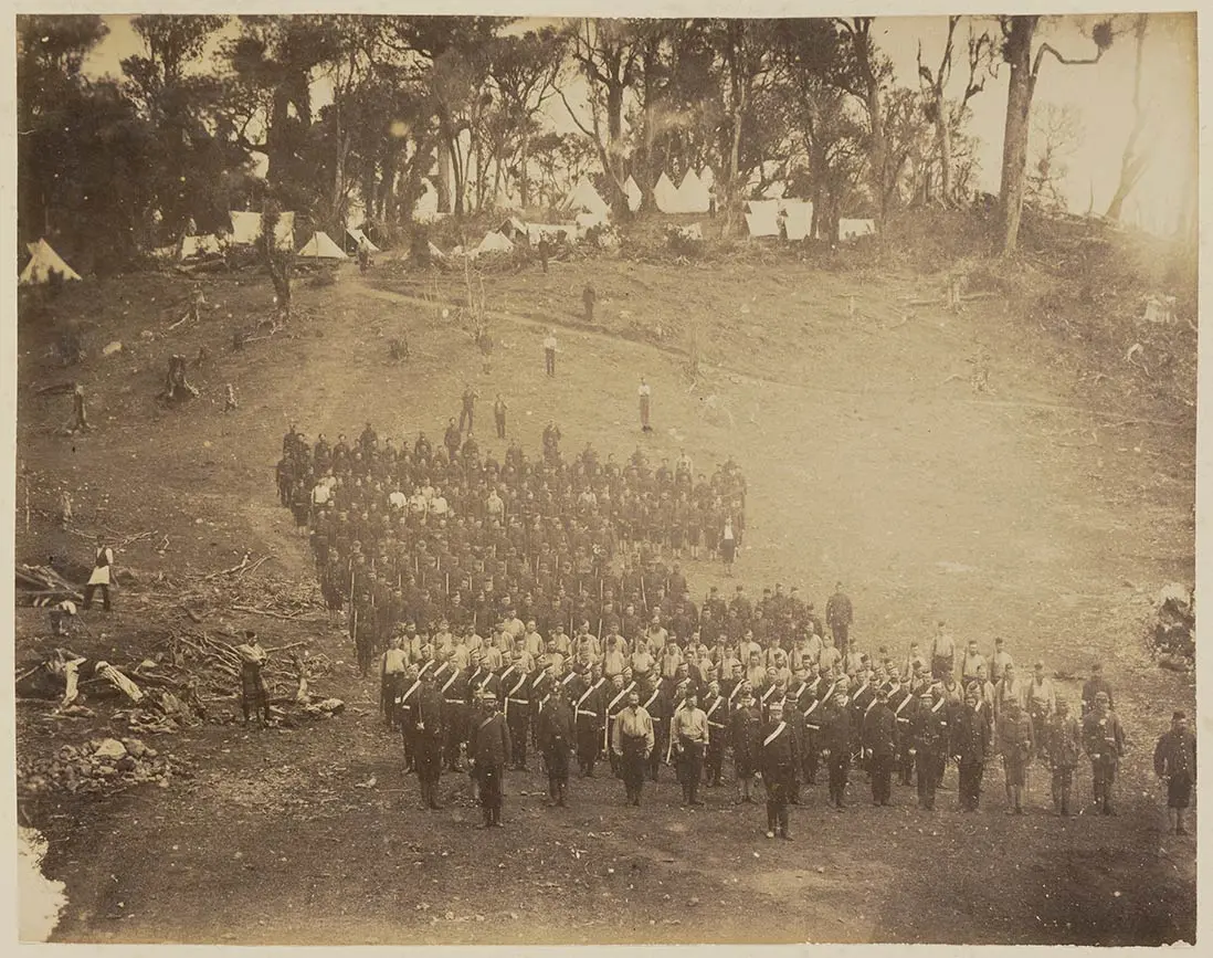 Black and white photo of a military camp at Parihaka showing government troops in uniform, standing in line formation.