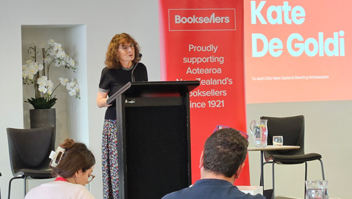 Kate De Goldi speaks at a podium at a Booksellers NZ event, with a red banner and projected slide displaying her name behind her.