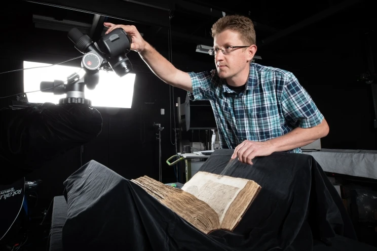 An imaging technician operates a camera on tripod that's angled so as to best capture the page of a partially opened bound volume.