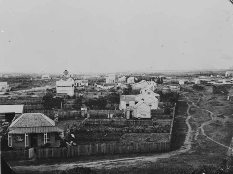 A black and white landscape photo showing newly built wooden houses with picket fences along a dirt road.