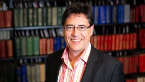 A formal portrait of a man with a bookshelf in the background. 