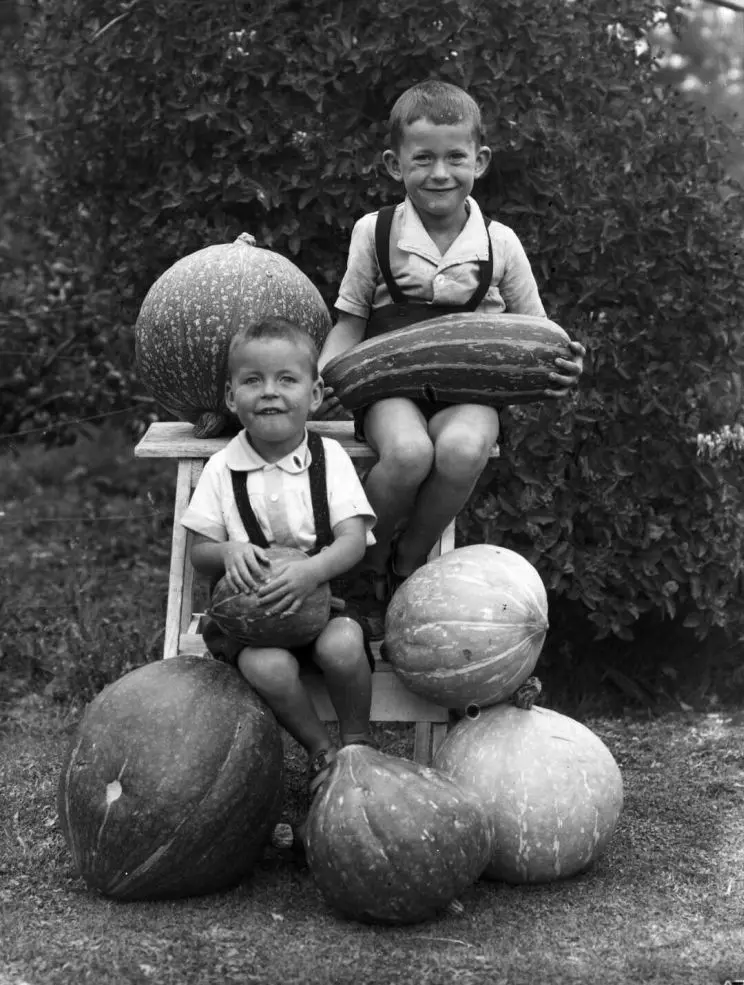 A black and white portrait of two young boys sitting surrounded by huge marrows and pumpkins.