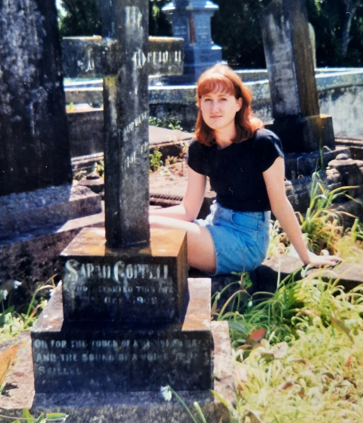 A colour photo of a young woman seated before a headstone in a graveyard.