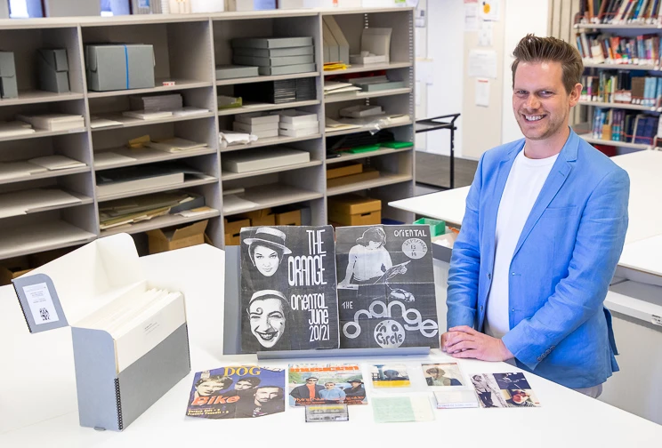 A man stands beside a table on which there are a range of printed items displayed along with an archival box containing white folders.