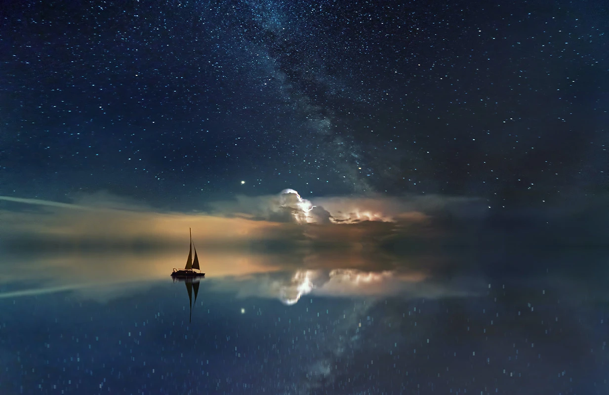 Colour photograph showing a boat sailing at sunset under the stars at night, and reflections on still water.
