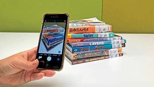 A hand holding a phone and taking a photo of a stack of books.