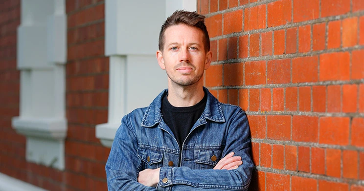 A man wearing a blue denim jacket and black t-shirt leaning on a brick wall.