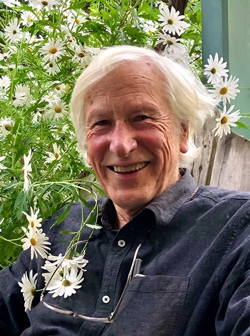 A smiling man siting in front of a plant with white flowers.