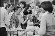 A black and white photo of three women in a market assessing a canned item. 